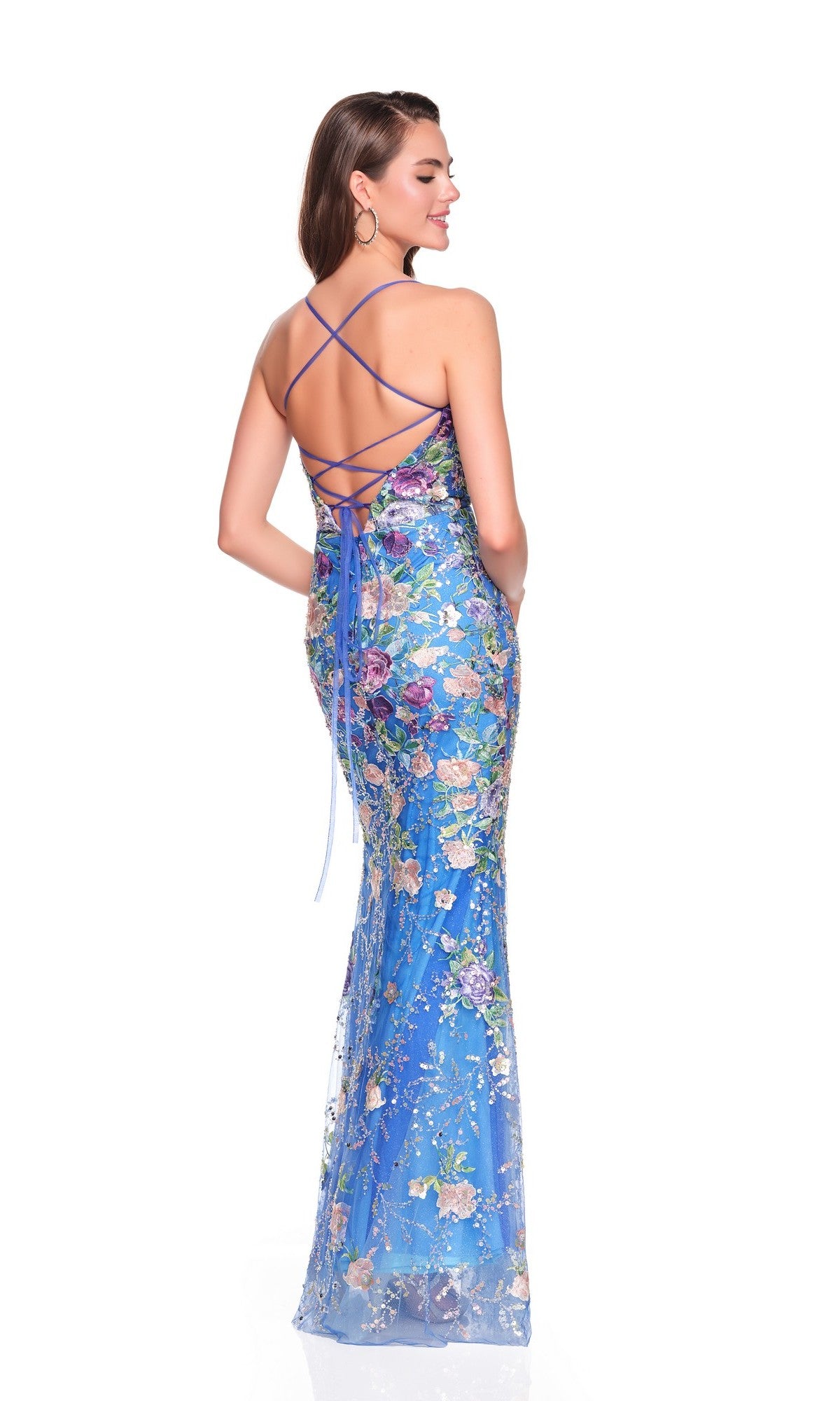A woman models the Dave and Johnny 12760 long prom dress in blue, featuring floral motifs and a lace-up open back, standing with hands in pockets and gazing to the side against a white backdrop.